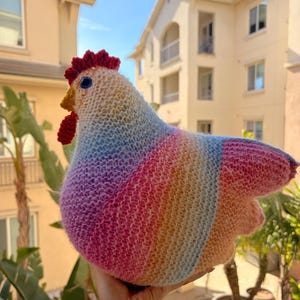 Puede incluir: Un pollo tejido a mano en un arcoíris de colores. El pollo tiene una cresta y una barbilla rojas, un pico amarillo y ojos azules. El cuerpo es un degradado de rosa, naranja, amarillo, azul y morado. Un artículo decorativo único.