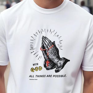 Op de afbeelding: Wit t-shirt met een afbeelding van biddende handen, een rozenkrans en een kruis. Het ontwerp bevat de woorden "WITH GOD" in het geel en "ALL THINGS ARE POSSIBLE" met het bijbelvers Mattheüs 19:26.