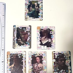 Può includere: Una collezione di sei biglietti fotografici decorativi. Ogni biglietto presenta un ritratto incorniciato con adesivi colorati, tra cui personaggi dei cartoni animati e forme pastello. Le immagini sembrano essere di un uomo in varie pose.