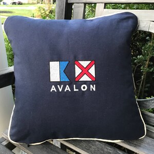 Può includere: Un cuscino blu navy con un designo di bandiera nautica bianca e rossa e la parola "AVALON" ricamata in bianco.