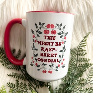May include: White ceramic mug with a red rim and handle. The mug has a design of red raspberries and green leaves with the text "This might be rasp-berry cordial" in red and gray lettering.