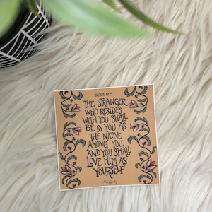 May include: A square tan sticker featuring black hand-lettered text that reads: Leviticus 19:34, The stranger who resides with you shall be to you as the native among you, and you shall love him as yourself. The text is framed by floral vines.