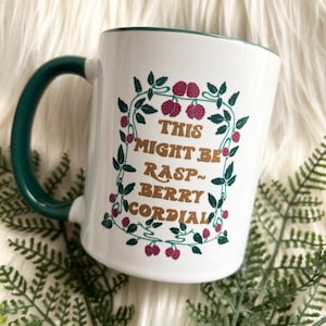 May include: White ceramic mug with a green rim and handle. The mug has a graphic of a raspberry vine with the text "This might be rasp-berry cordial" in gold lettering.