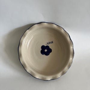 Op de afbeelding: Een ronde keramische schaal met een geschulpte rand en een crèmekleurige binnenkant. Het midden is voorzien van een donkerblauw geschilderd bloemmotief en de tekst .LOVE in donkerblauwe letters. De rand is afgewerkt met een dunne donkerblauwe lijn.
