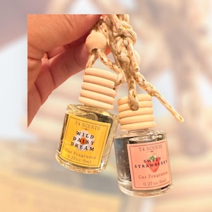 May include: Two small glass car fragrance bottles with wooden caps and braided hanging cords. One bottle has a yellow label reading T4 SCENTS WILD DAISY DREAM 0.27oz/8ml, and the other has a pink label reading T4 SCENTS SWEET STRAWBERRY 0.27oz/8ml.