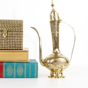 May include: A vintage brass pitcher with an elongated spout and ornate floral engravings. Beside it, a small gold-studded box and a stack of books with colorful covers. The pitcher is approximately 12 inches tall.