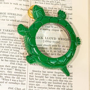 May include: A green turtle-shaped magnifying glass with a circular lens in the center of its shell. The turtle has a glossy finish, four legs, a head, and a tail. A small gold-colored metal bell is attached to the top of the turtle's head.