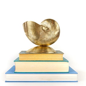 May include: A golden, nautilus-shaped sculpture sits atop a stack of books. The sculpture has a textured surface and is balanced on a small, round base. The books have blue and teal covers with gold-edged pages, creating a decorative display.