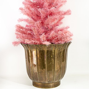 May include: A vibrant pink artificial Christmas tree with needle-like branches is set inside a large, antique-style brass planter. The planter features a distinct vertical ribbed design with a wide, flared rim and a solid, circular base.