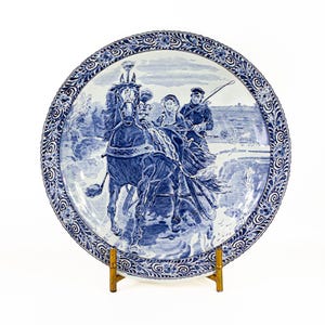 May include: A blue and white decorative plate featuring a horse-drawn carriage scene. The plate has a detailed border with floral and scroll designs. The image depicts a horse pulling a carriage with two people and a driver.