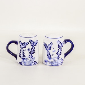 May include: A pair of ceramic salt and pepper shakers. Each shaker is white with a blue floral design and a dark blue handle. The shakers have a rounded top with several small holes for dispensing.