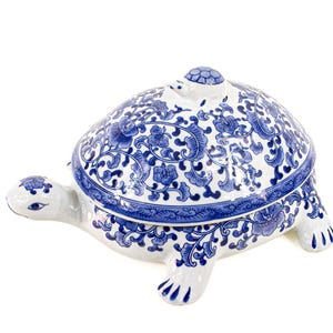 May include: A white ceramic turtle-shaped lidded container featuring a blue floral pattern across the shell, head, and legs. The turtle has a small turtle figure on top of its shell, a pointed head with painted eyes, and four distinct feet.