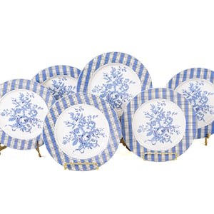 May include: A set of six white ceramic plates with a blue floral design in the center. The plates have a blue and white plaid border. Each plate is displayed on a gold-colored metal stand.