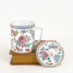 May include: White ceramic mug and lid with a floral design. The mug features a handle and is decorated with pink, red, orange, and blue flowers. The lid has a matching floral pattern and a small knob. The mug and lid are placed on a vintage book.