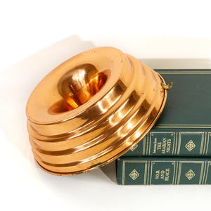 May include: A copper-colored bundt pan with a fluted design. The pan has a central tube and a small handle. It is resting on top of two green books with gold lettering.