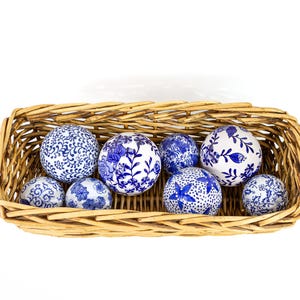 May include: A rectangular woven basket filled with decorative spheres. The spheres feature intricate blue and white floral patterns. The basket is made of light brown woven material, and the spheres vary in size.