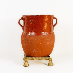 May include: A rustic, terracotta-colored ceramic vase with two loop handles. The vase sits on a decorative brass stand with a square top and tassel-shaped feet. The vase has a glossy, reddish-brown upper section and a matte, lighter brown lower section.