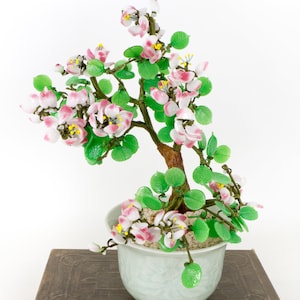 May include: A decorative glass bonsai tree with pink and white flowers, green leaves, and a brown trunk, potted in a light blue ceramic pot. The bonsai is set on a vintage book with a worn, brown cover.