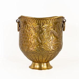 May include: A polished brass bucket featuring an embossed design of grape clusters and vine leaves on the exterior. The vessel has a flared rim with a rope-like texture, a pedestal base, and two circular ring handles attached to the sides.