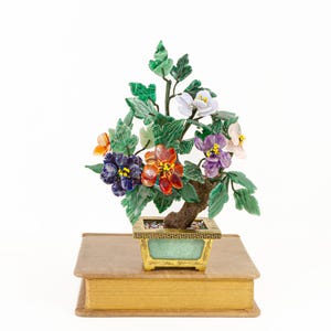 May include: A colorful bonsai tree sculpture with green leaves and vibrant flowers in shades of orange, purple, white, and blue. The tree is in a gold-trimmed pot, resting on a gold-edged book.