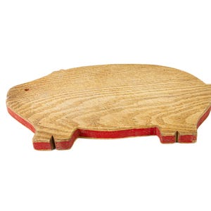 May include: Wooden pig-shaped cutting board with a red painted border. The board has a natural wood grain pattern and a small hole near the top. The pig-shaped design adds a playful touch to this kitchen item.
