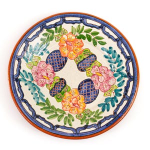 May include: A hand-painted ceramic plate with a floral design. The plate features a white center with pink and orange flowers, blue lattice patterns, and green leaves. The rim has a blue and brown border.