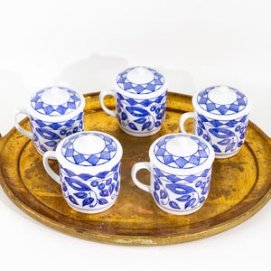 May include: Five white ceramic mugs with blue floral and geometric designs, each with a matching lid. The mugs are arranged on a gold-toned tray, creating a cohesive set. The handles are small and curved.