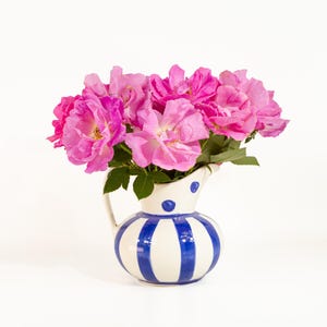 May include: A ceramic pitcher with a white base and vertical blue stripes, featuring a handle and a spout decorated with blue dots. The pitcher is filled with a bouquet of vibrant pink roses with green leaves, creating a colorful floral display.