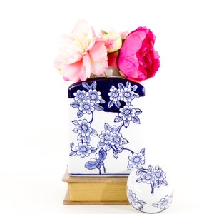 May include: A white and blue floral ceramic vase with a dark blue rim, holding pink and white flowers. A matching small round decorative piece sits beside the vase. The vase and decorative piece are on a book.