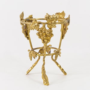 May include: A gold-colored metal decorative stand with a three-legged base. The stand features a round top and is adorned with grape and leaf designs. The stand is likely used for displaying small items.