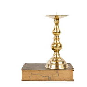 May include: A gold-colored candlestick holder with a flared top and ornate stem, resting on a worn, brown book. The candlestick is designed to hold a single candle. The background is plain white.