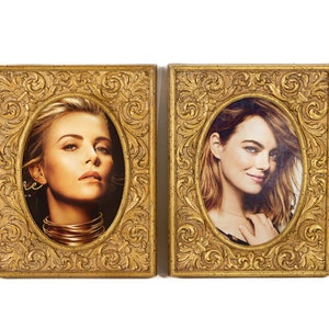 May include: Two rectangular picture frames with ornate, raised gold-colored scrollwork and floral detailing. Each frame features an oval-shaped opening containing a portrait photograph of a person. The frames are positioned side-by-side against a white background.