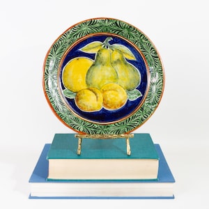 May include: A decorative ceramic plate featuring a fruit design of pears, peaches, and a lemon on a blue background. The plate has a green and red border. The plate is displayed on a gold stand, atop two books.