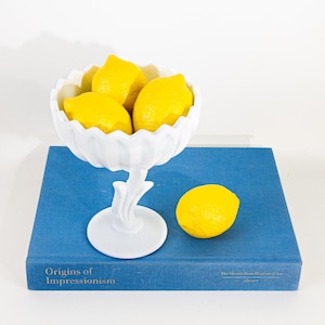May include: A white, scalloped-edge compote filled with bright yellow lemons, resting on a blue book. One lemon sits beside the compote. The book has the title "Origins of Impressionism" printed on the cover.
