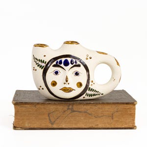 May include: A cream-colored ceramic vessel with two top openings and a side handle. The front features a hand-painted face framed by a dark circle, with blue eyes, dark eyebrows, and gold-toned lips. Green fern-like patterns decorate the sides.