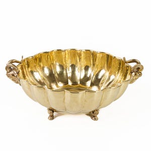 May include: A gold-colored decorative bowl with a scalloped rim and three small feet. The bowl has two handles with ram's head designs. The interior of the bowl is highly reflective, and the overall design is ornate.