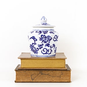 May include: A white and blue ceramic jar with a lid, featuring a floral design. The jar is placed on top of two stacked, aged books with gold-edged pages. The background is plain white.