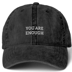May include: A black baseball cap with a distressed, washed-out finish. The front of the cap features the embroidered white text "YOU ARE ENOUGH" in a clean, sans-serif font. The hat has a curved brim and a structured six-panel crown design.