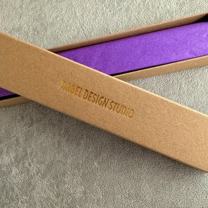 A long, narrow, empty kraft jewelry box with the words Isabel Design Studio embossed in gold letters.