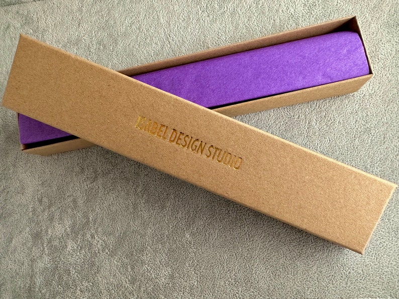 A long, narrow, empty kraft jewelry box with the words Isabel Design Studio embossed in gold letters.