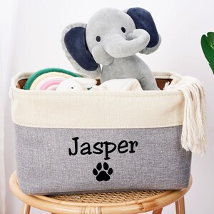 May include: A gray and white storage bin with a white tassel. The bin has the name "Jasper" and a paw print embroidered on the front. The bin is filled with toys, including a gray elephant stuffed animal and a rainbow-colored toy.