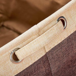 May include: Close-up of a beige and brown fabric storage bin with a drawstring closure. The bin has two metal grommets on the top edge.