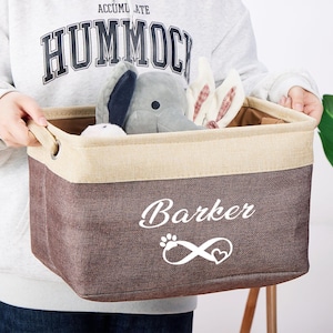 May include: A brown and beige canvas storage bin with the word "Barker" and an infinity symbol with a heart in the center printed in white. The bin is filled with stuffed animals, including a gray elephant and two white bunnies.