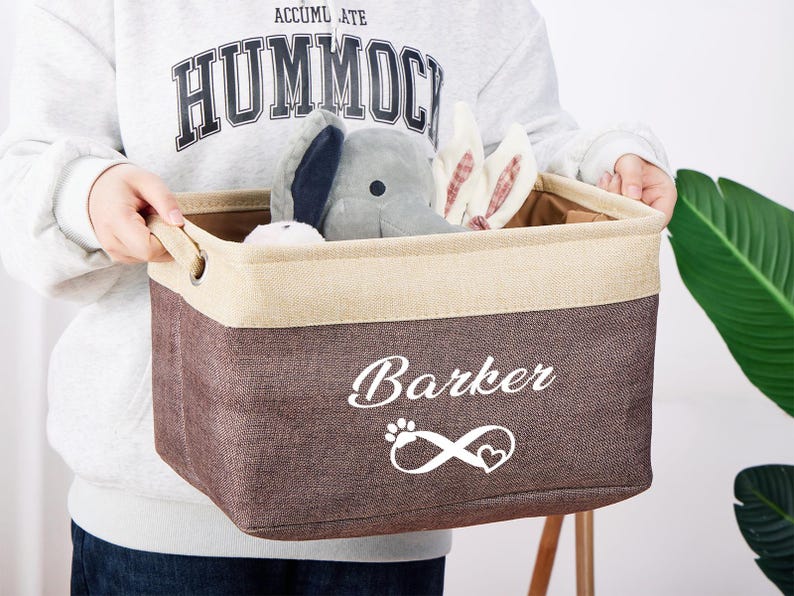 May include: A brown and beige canvas storage bin with the word "Barker" and an infinity symbol with a heart in the center printed in white. The bin is filled with stuffed animals, including a gray elephant and two white bunnies.