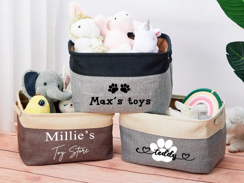 May include: Three gray canvas storage bins with black trim and paw print designs. The bins are labeled "Max's toys", "Millie's Toy Store", and "teddy".