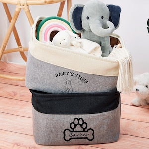May include: Two gray fabric storage bins with black trim. The top bin has the text "Daisy's Stuff" and a small cat illustration. The bottom bin has a paw print and the text "Barker". The bins are filled with toys, including a rainbow ring, a stuffed elephant, and a stuffed dog.