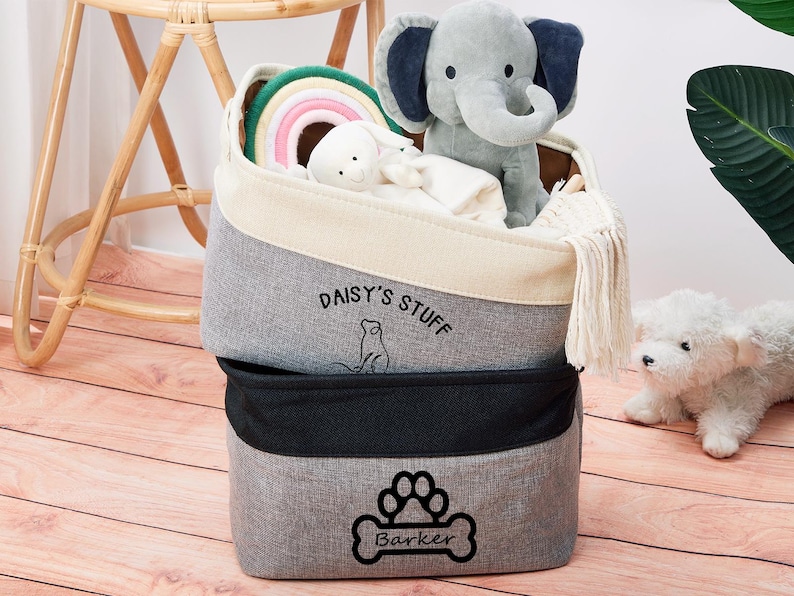 May include: Two gray fabric storage bins with black trim. The top bin has the text "Daisy's Stuff" and a small cat illustration. The bottom bin has a paw print and the text "Barker". The bins are filled with toys, including a rainbow ring, a stuffed elephant, and a stuffed dog.