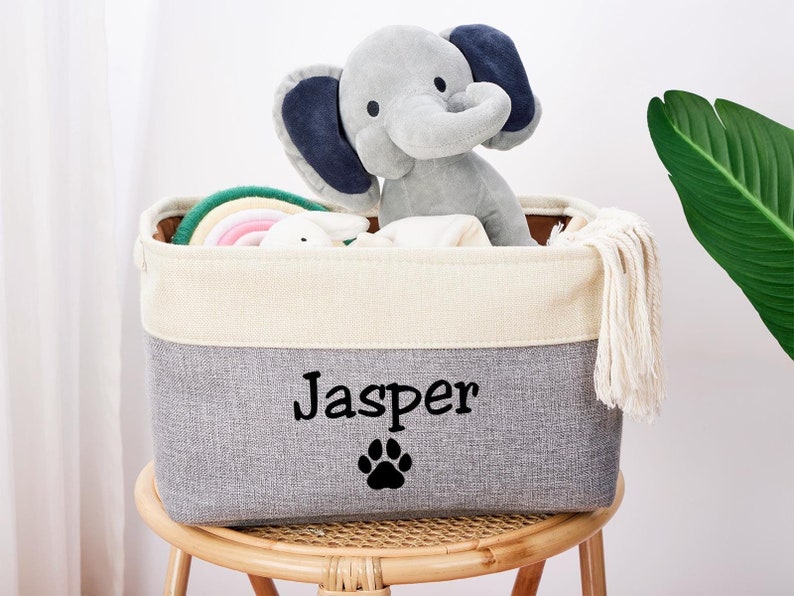 May include: A gray and white storage bin with a white tassel. The bin has the name "Jasper" and a paw print embroidered on the front. The bin is filled with toys, including a gray elephant stuffed animal and a rainbow-colored toy.