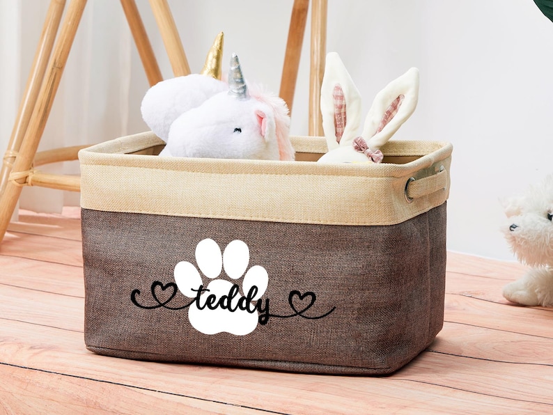 May include: A brown and beige fabric storage bin with a white paw print and the text "teddy" in black. The bin is filled with a white unicorn and a white bunny plush toy.