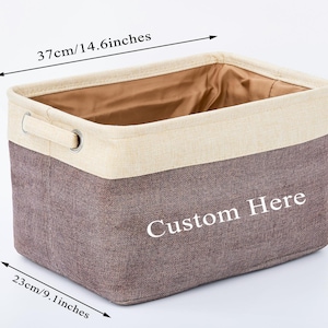 May include: A brown and beige fabric storage bin with a white handle and the text "Custom Here" printed on the front. The bin measures 14.6 inches wide, 10.6 inches tall, and 9 inches deep.